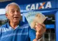 Idoso brasileiro segura cédulas de real em frente a uma agência da Caixa, representando a consulta aos depósitos do INSS de até R$ 8.475,55.