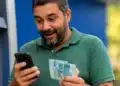 Contribuinte consulta no celular o segundo lote da restituição do Imposto de Renda 2026 enquanto segura cédulas de real.