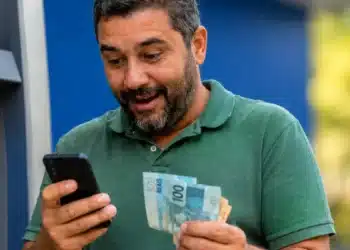 Contribuinte consulta no celular o segundo lote da restituição do Imposto de Renda 2026 enquanto segura cédulas de real.