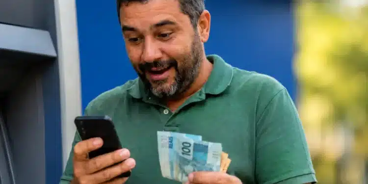 Contribuinte consulta no celular o segundo lote da restituição do Imposto de Renda 2026 enquanto segura cédulas de real.