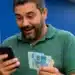 Contribuinte consulta no celular o segundo lote da restituição do Imposto de Renda 2026 enquanto segura cédulas de real.