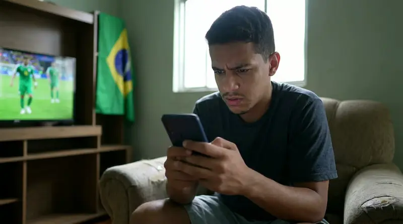 Jovem observa celular com expressão preocupada enquanto acompanha jogo de futebol na TV, em alerta contra golpes ligados à Copa.