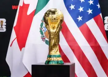 Troféu da Copa do Mundo da FIFA 2026 em destaque com bandeiras dos países-sede ao fundo durante fase eliminatória.