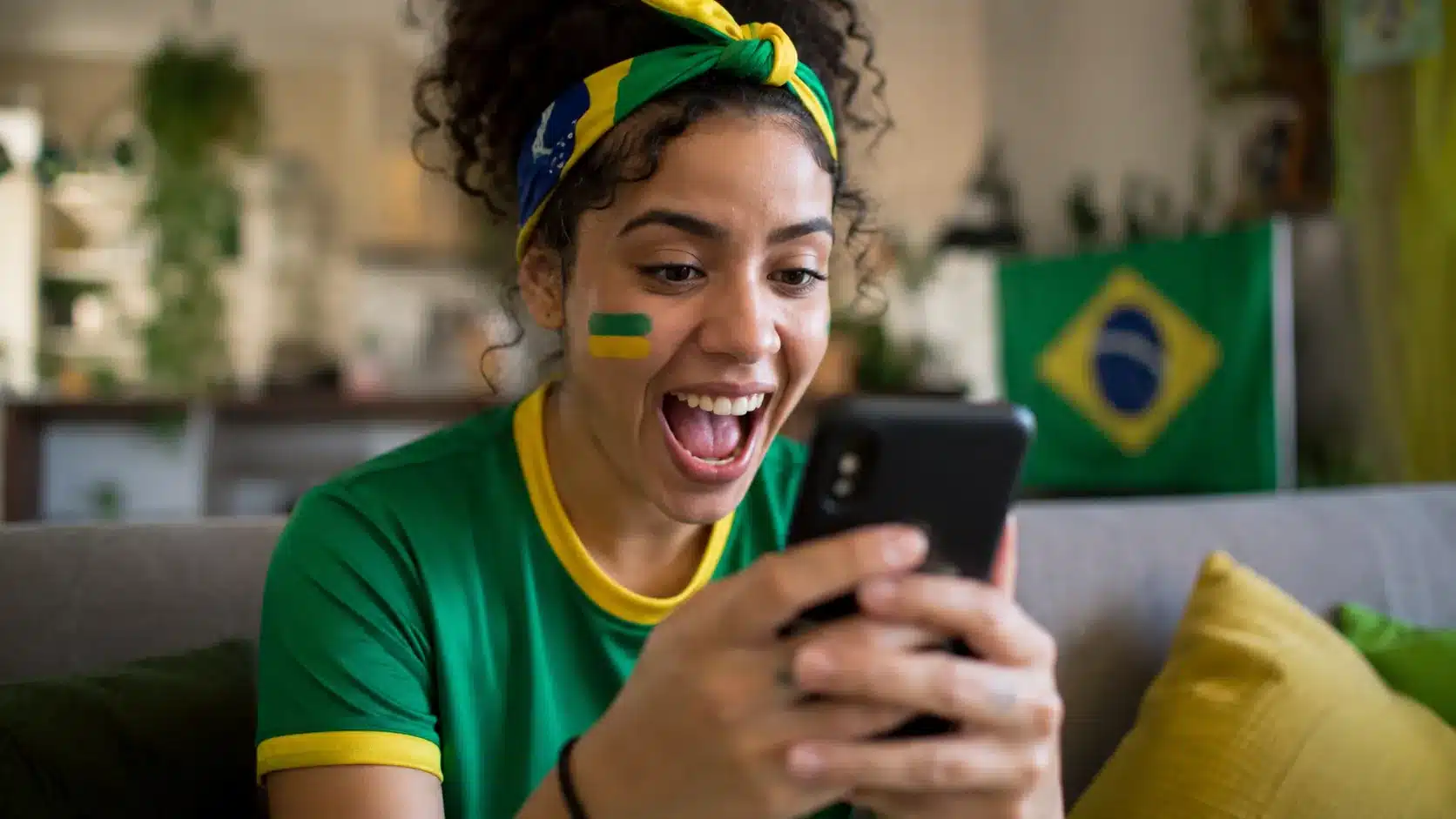 Jovem torcedora brasileira animada segurando smartphone, interagindo com figurinhas e filtros da Copa do Mundo no WhatsApp, com pintura facial verde e amarela e faixa de torcida.
