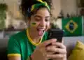 Jovem torcedora brasileira animada segurando smartphone, interagindo com figurinhas e filtros da Copa do Mundo no WhatsApp, com pintura facial verde e amarela e faixa de torcida.
