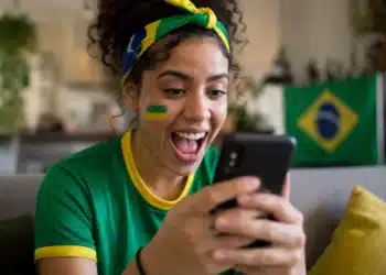Jovem torcedora brasileira animada segurando smartphone, interagindo com figurinhas e filtros da Copa do Mundo no WhatsApp, com pintura facial verde e amarela e faixa de torcida.
