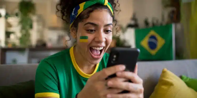 Jovem torcedora brasileira animada segurando smartphone, interagindo com figurinhas e filtros da Copa do Mundo no WhatsApp, com pintura facial verde e amarela e faixa de torcida.