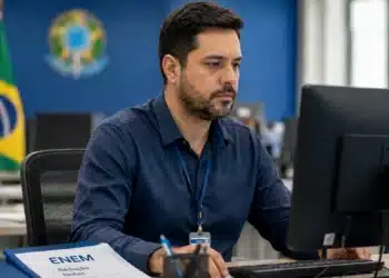 Corretor de redação analisa informações no computador enquanto uma pasta do Enem sobre redação e notas aparece sobre a mesa.
