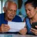 Idoso e mulher de meia-idade conferindo documento e celular em casa, com nota de R$ 100, representando os dois grupos que ainda aguardam o pagamento da segunda parcela do 13º do INSS