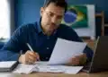 Homem brasileiro revisando documentos do Imposto de Renda 2026 enquanto utiliza o computador, representando a correção de dados enviados à Receita Federal.