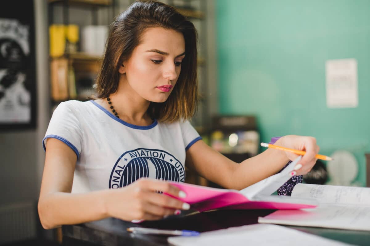 Jovem concentrada estuda com lápis na mão enquanto folheia cadernos e anotações sobre a mesa