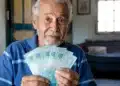 Aposentado brasileiro segurando notas de cem reais em casa, representando os depósitos de R$ 8.475,55 do INSS nesta terça-feira, 2 de junho.