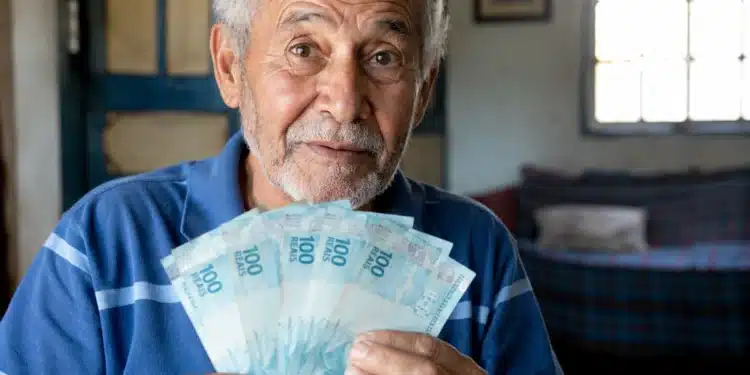 Aposentado brasileiro segurando notas de cem reais em casa, representando os depósitos de R$ 8.475,55 do INSS nesta terça-feira, 2 de junho.