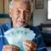 Aposentado brasileiro segurando notas de cem reais em casa, representando os depósitos de R$ 8.475,55 do INSS nesta terça-feira, 2 de junho.