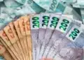 Notas de dinheiro real brasileiro sendo seguradas, com notas de R$ 100 ao fundo, ilustrando os depósitos de até R$ 2.625 da Caixa Econômica Federal.