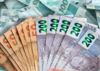 Notas de dinheiro real brasileiro sendo seguradas, com notas de R$ 100 ao fundo, ilustrando os depósitos de até R$ 2.625 da Caixa Econômica Federal.