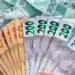 Notas de dinheiro real brasileiro sendo seguradas, com notas de R$ 100 ao fundo, ilustrando os depósitos de até R$ 2.625 da Caixa Econômica Federal.