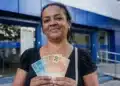 Mulher brasileira segurando cédulas de real em frente a uma agência da Caixa Econômica Federal, representando os depósitos de R$ 2.900 na quinta-feira, 25 de junho.