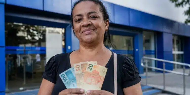 Mulher brasileira segurando cédulas de real em frente a uma agência da Caixa Econômica Federal, representando os depósitos de R$ 2.900 na quinta-feira, 25 de junho.