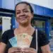 Mulher brasileira segurando cédulas de real em frente a uma agência da Caixa Econômica Federal, representando os depósitos de R$ 2.900 na quinta-feira, 25 de junho.