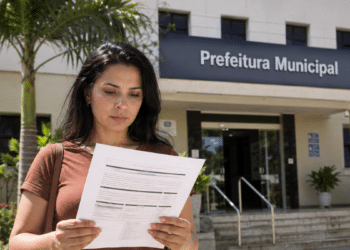 Mulher lê documento de concurso público em frente a uma prefeitura municipal.