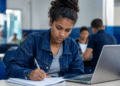 Jovem mulher concentrada escrevendo em caderno ao lado de laptop em sala de aula.