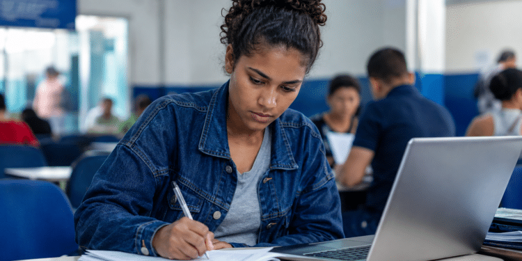 Jovem mulher concentrada escrevendo em caderno ao lado de laptop em sala de aula.