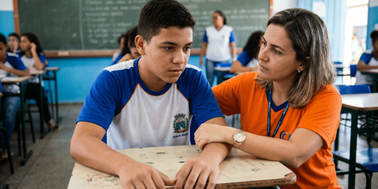 Estudante uniformizado recebe apoio de profissional em sala de aula durante atividade escolar