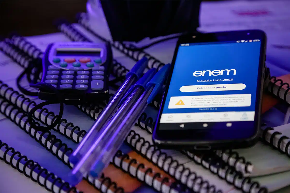 Celular com tela do Enem sobre cadernos e materiais escolares, representando o novo prazo para atendimento especializado e nome social em 2026.