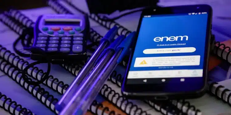 Celular com tela do Enem sobre cadernos e materiais escolares, representando o novo prazo para atendimento especializado e nome social em 2026.