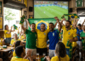 Torcedores vibram com a bandeira do Brasil diante de um telão durante jogo da Seleção em um bar
