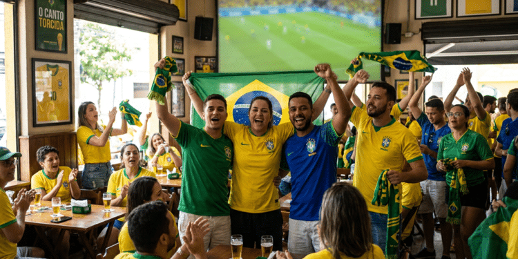 Torcedores vibram com a bandeira do Brasil diante de um telão durante jogo da Seleção em um bar