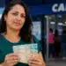 Mulher segurando cédulas de dinheiro em frente a uma agência da Caixa, simbolizando a liberação do FGTS no valor de R$ 900 disponível até 30 de junho