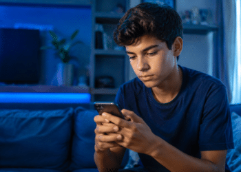 Jovem de cabelos escuros e camiseta azul marinho usa celular em ambiente noturno com iluminação LED azul.