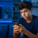 Jovem de cabelos escuros e camiseta azul marinho usa celular em ambiente noturno com iluminação LED azul.