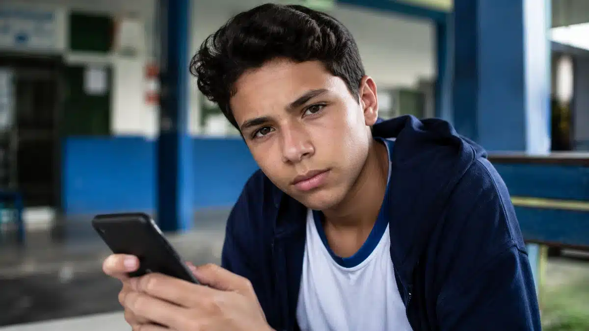 Jovem estudante em escola pública consulta celular com expressão preocupada, representando o possível bloqueio do Pé-de-Meia por baixa frequência.