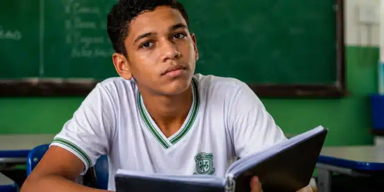 Estudante brasileiro com caderno em sala de aula pública, representando regra de frequência para recebimento do benefício mensal do Pé-de-Meia.