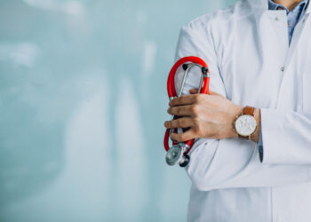 Médico de jaleco branco segura estetoscópio vermelho com os braços cruzados