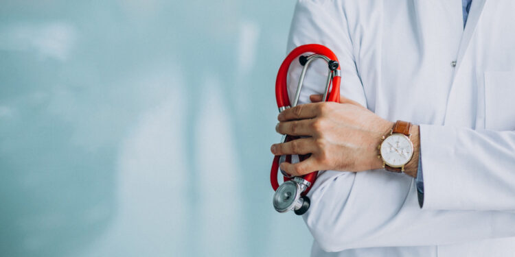 Médico de jaleco branco segura estetoscópio vermelho com os braços cruzados