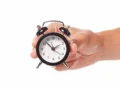 Mão segurando relógio analógico em referência ao uso correto da abreviatura de horas e minutos.