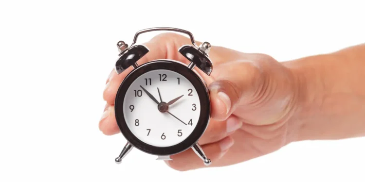 Mão segurando relógio analógico em referência ao uso correto da abreviatura de horas e minutos.