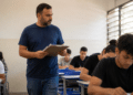 Professor com barba e camiseta azul vigia estudantes concentrados fazendo prova em sala iluminada.