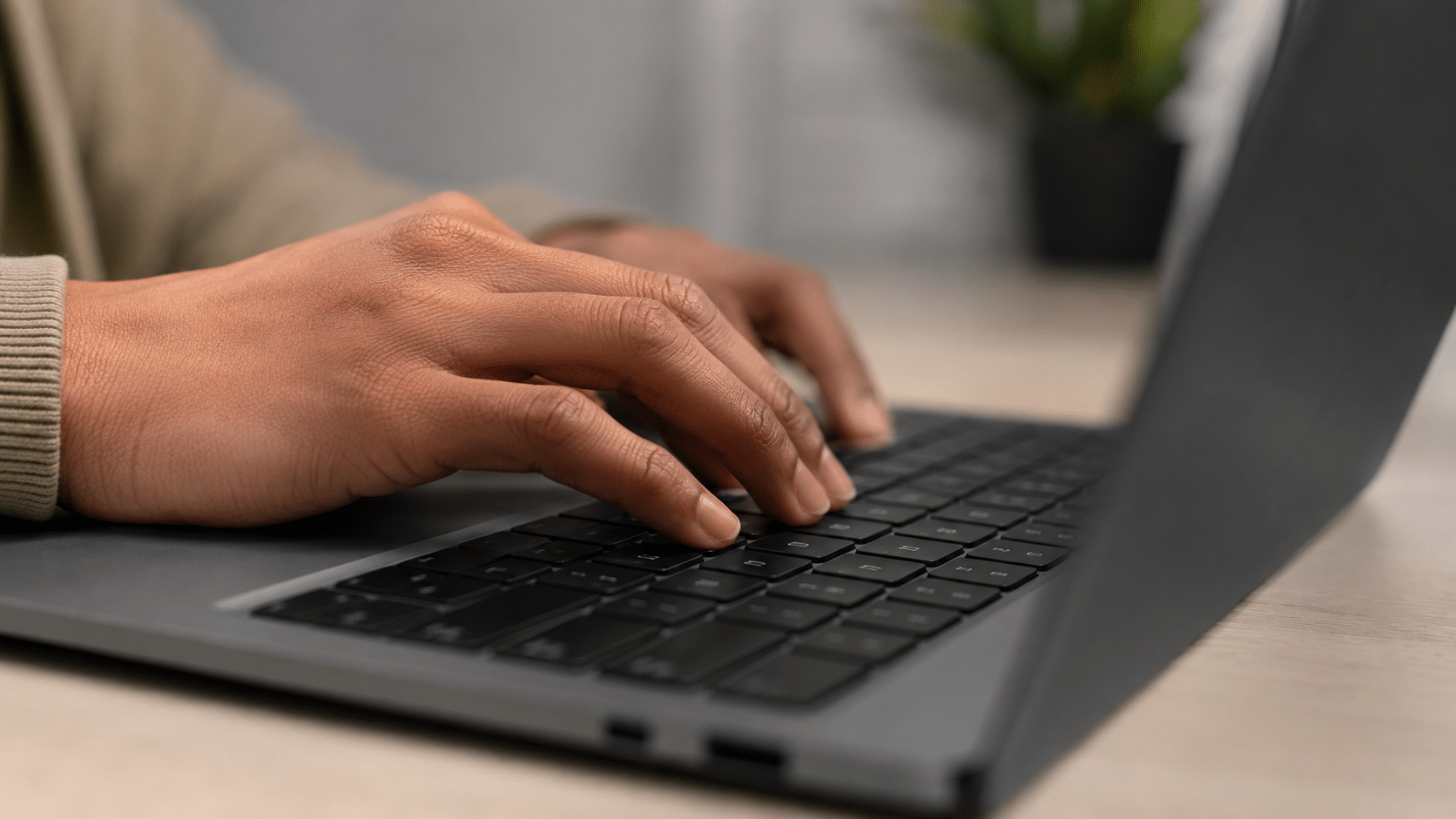 Mãos morenas digitando em teclado de notebook preto sobre mesa clara com planta ao fundo.