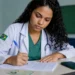 Médica brasileira escrevendo em uma sala de aula, com celular e documento sobre a mesa, representando a inscrição para o Enamed 2026.
