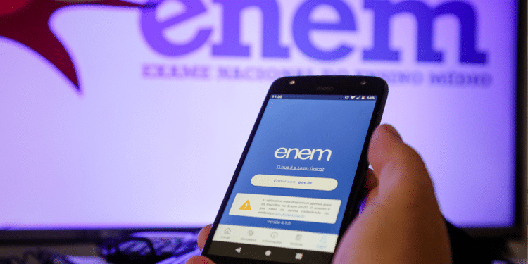 Mão segura celular com aplicativo Enem aberto diante de tela exibindo logo do Exame Nacional.