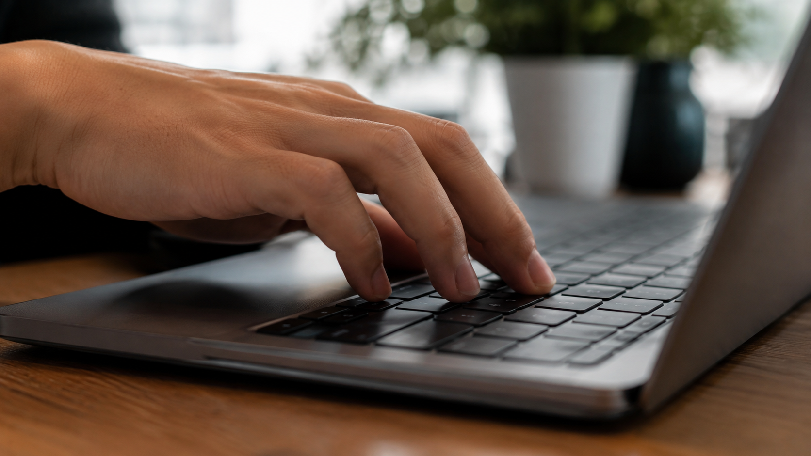 Mão de adulto digitando em teclado de notebook com vaso de planta desfocado ao fundo.