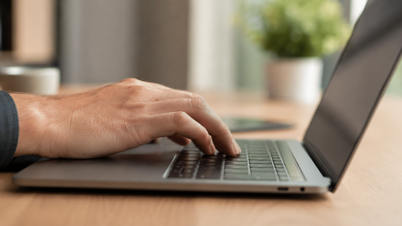 Mão masculina digitando em teclado de notebook prateado com fundo desfocado.