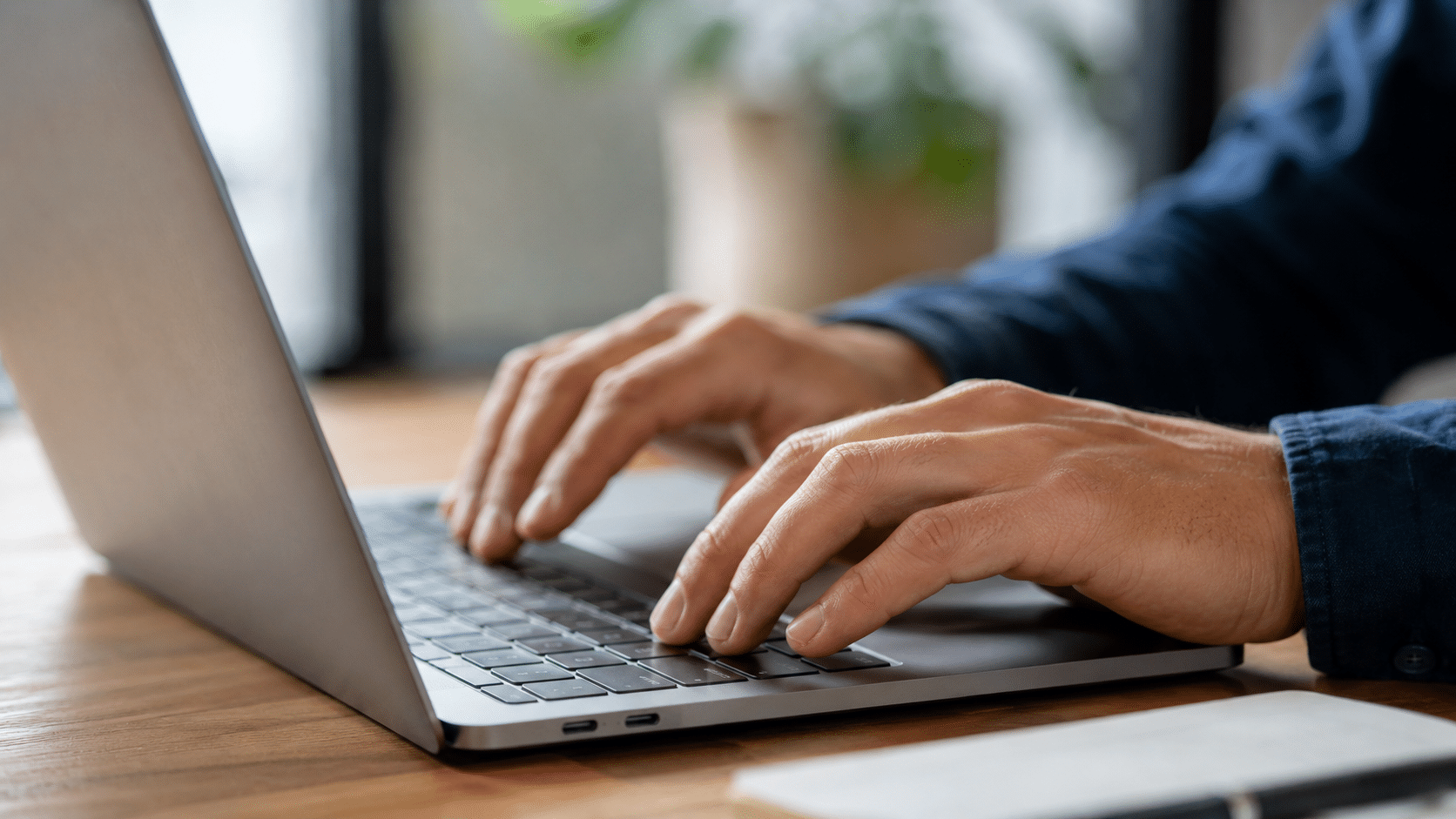 Inscrição Prefeitura Mãos masculinas digitando em notebook sobre mesa de madeira com planta ao fundo.