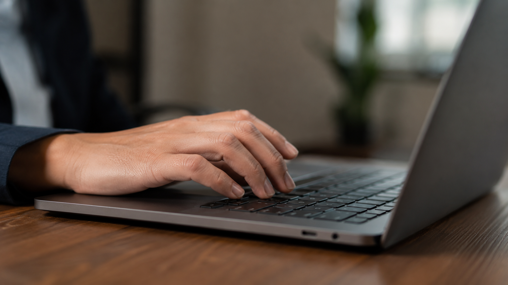 Mão masculina digitando em laptop sobre mesa de madeira com fundo desfocado.