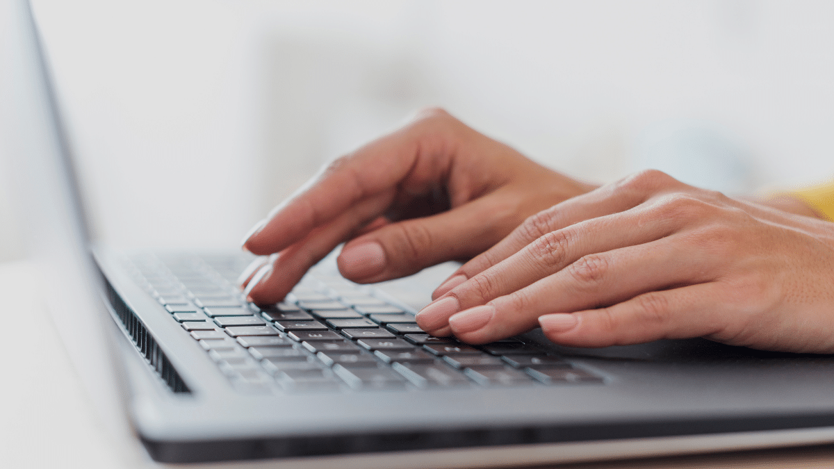 Mãos femininas digitando em teclado de notebook preto com fundo desfocado claro.